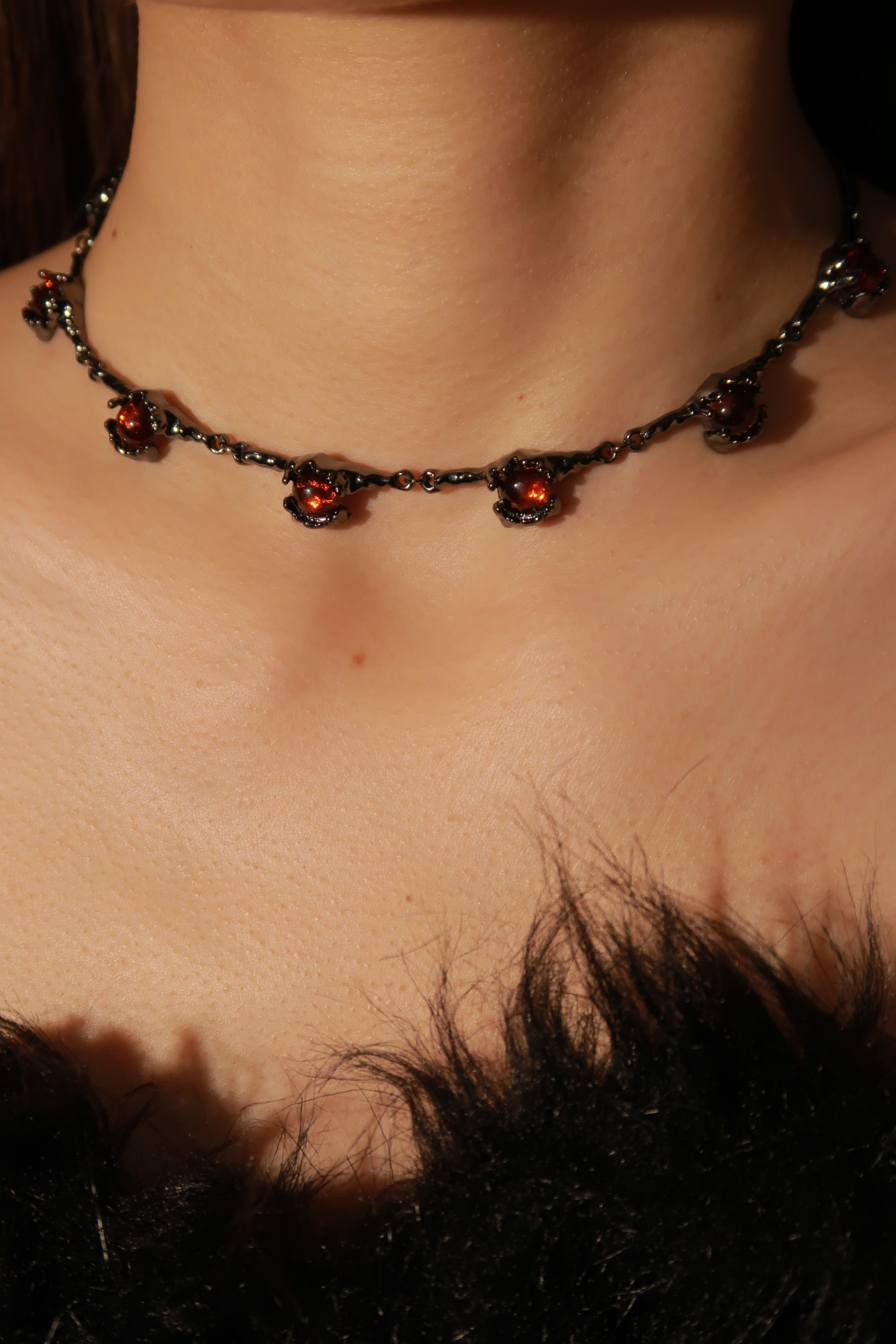 A close-up of a crimson rose choker necklace featuring red gemstones and black Chain detailing, worn by a person with black hair.