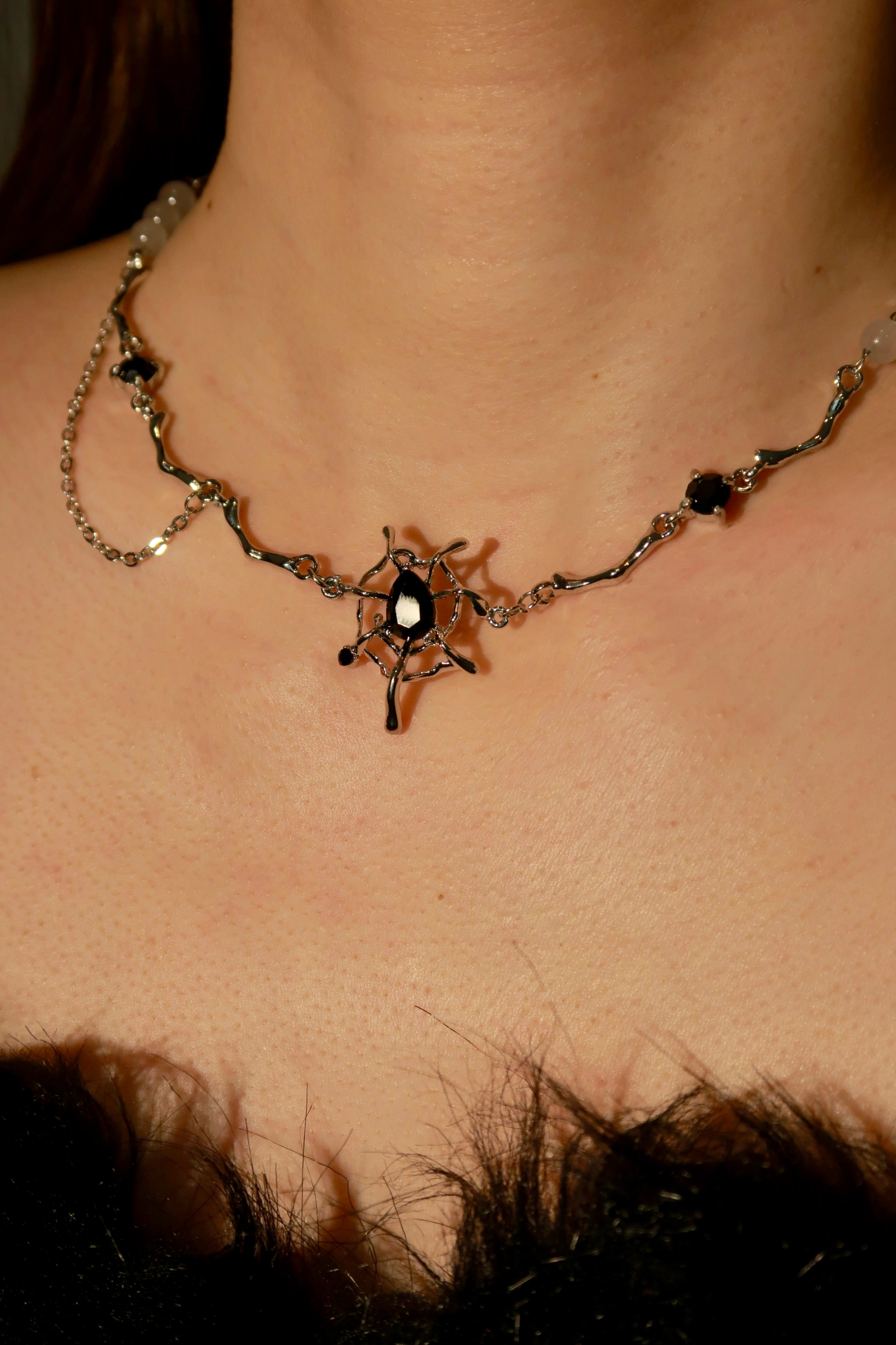 A close-up image of a necklace with a silver and black color scheme, featuring a central pendant that has a spider design, surrounded by small spherical charms linked by chain.