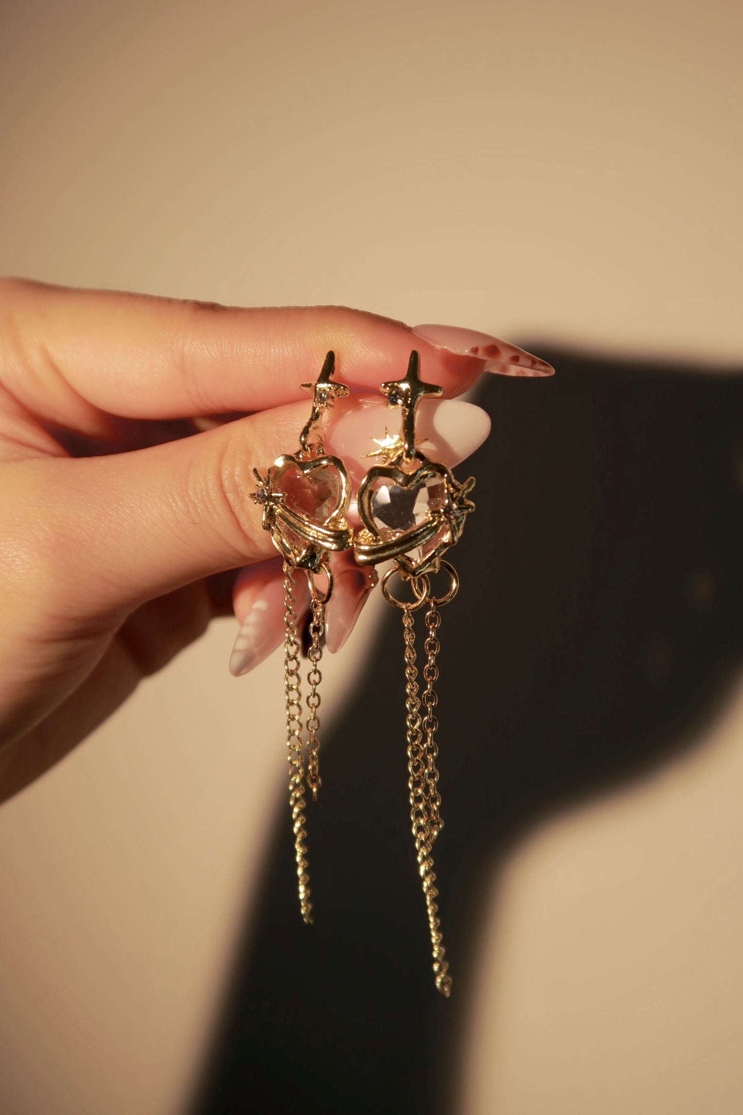 Gold earrings with chains held by a hand against a beige background