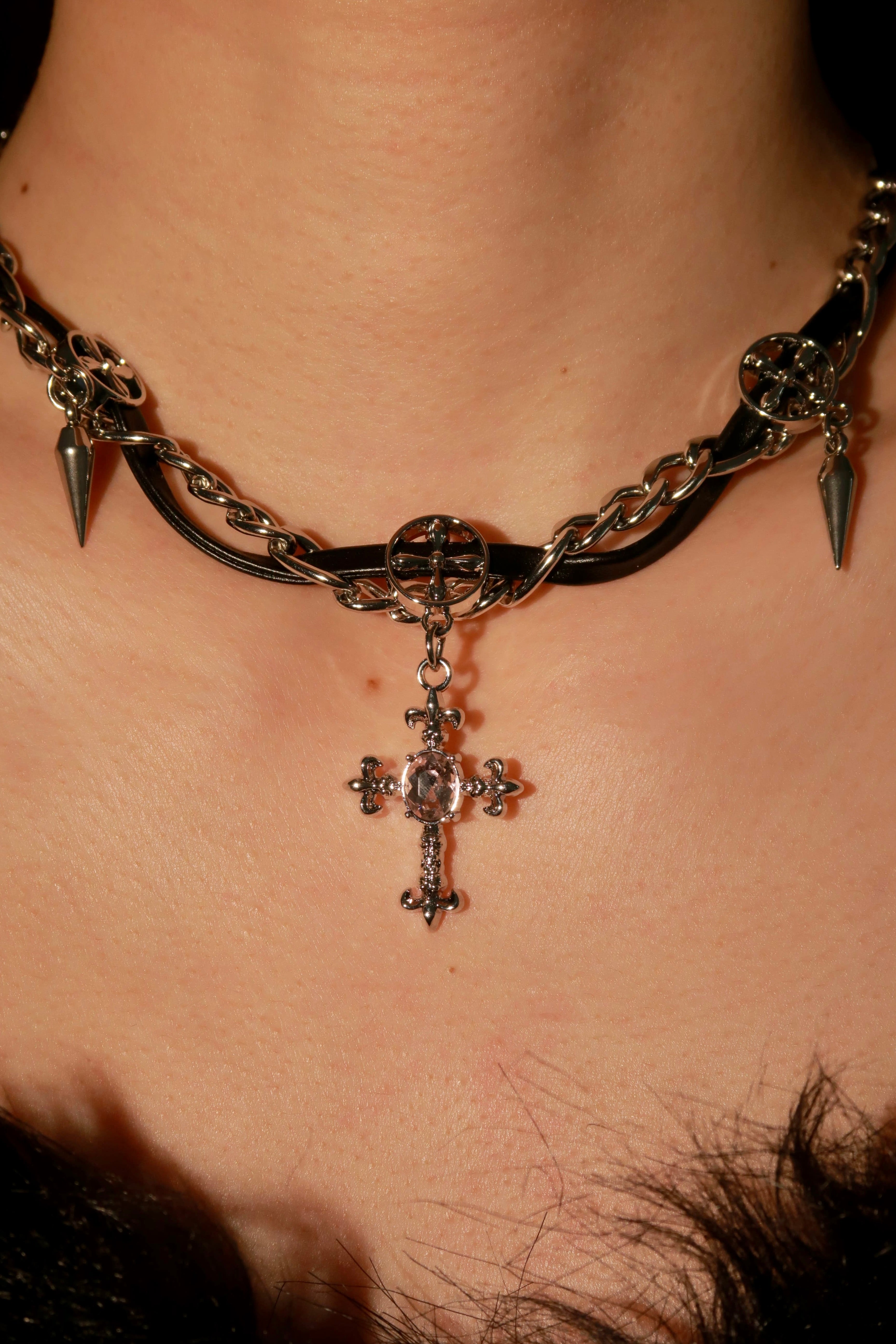 A close-up image of a Gothic Spike Cross Choker necklace featuring a black leather strap, metal chains, spikes, and a cross pendant.