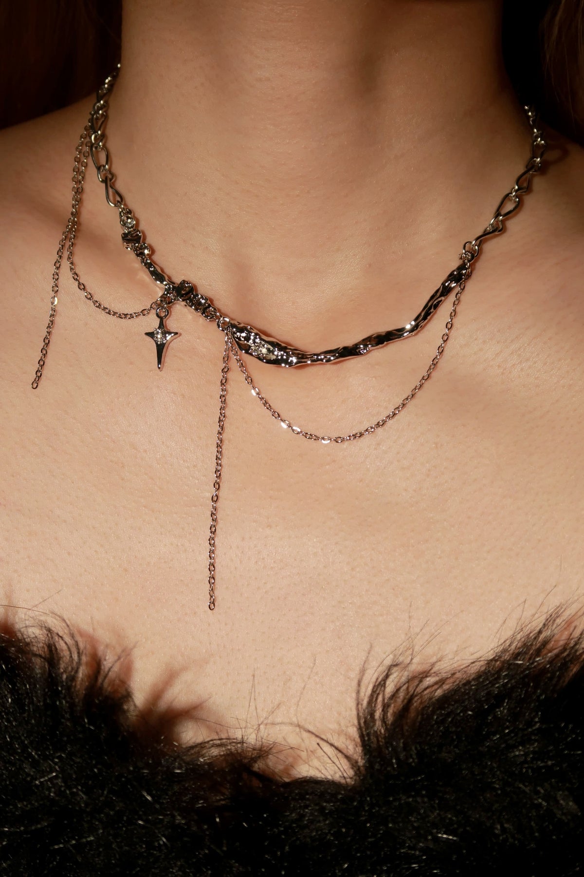 A close-up of a person's neck and upper chest wearing an ornate layered necklace with multiple chains, a twisted dark chain, a small cross pendant, and metallic details.