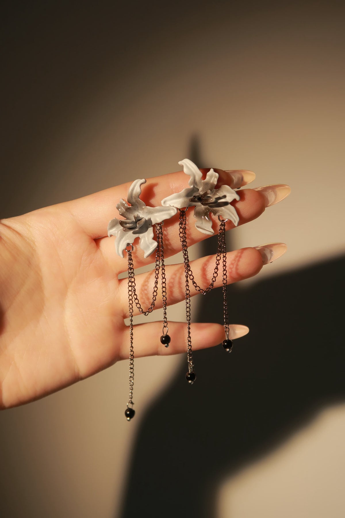 A hand holding a pair of dangling earrings with white flower-shaped tops, dark metal chains, and small black bead drops against a warm beige background.